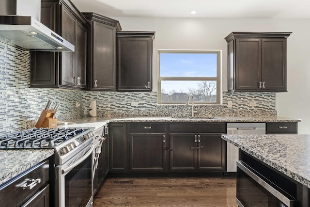 kitchen with appliances with stainless steel finishes, a sink, wall chimney range hood, dark brown cabinets, and light stone countertops