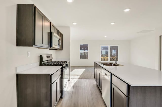 kitchen with stainless steel appliances, a sink, dark brown cabinets, light countertops, and an island with sink