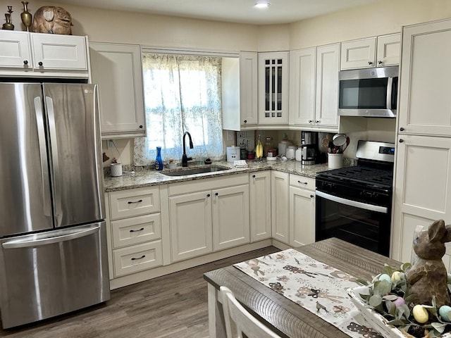 kitchen with stainless steel appliances, a sink, white cabinetry, light stone countertops, and glass insert cabinets