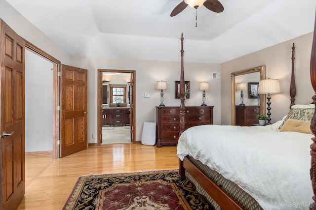 bedroom with ceiling fan, a raised ceiling, light wood-style flooring, and connected bathroom