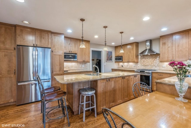 kitchen featuring light wood finished floors, wall chimney exhaust hood, a kitchen island with sink, stainless steel appliances, and a sink