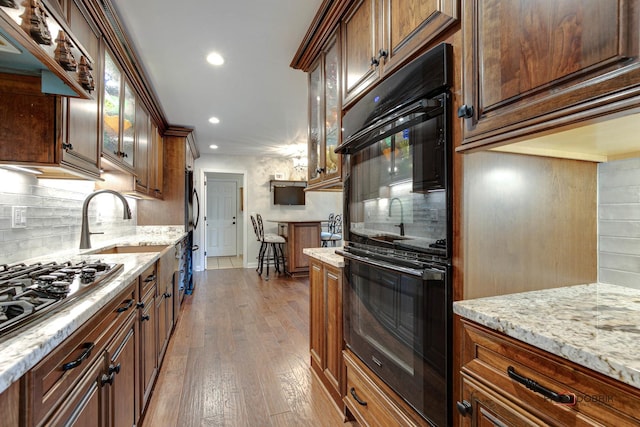 kitchen with dobule oven black, glass insert cabinets, wood finished floors, light stone countertops, and gas stovetop