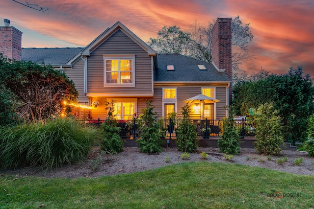 back of house at dusk with a shingled roof, a chimney, and a yard