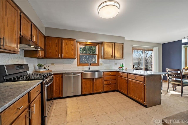 kitchen featuring stainless steel appliances, brown cabinetry, a sink, a peninsula, and under cabinet range hood