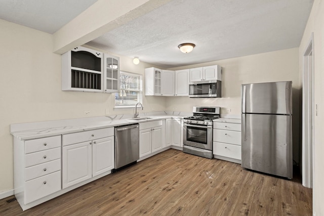 kitchen with appliances with stainless steel finishes, glass insert cabinets, white cabinets, a sink, and light wood-type flooring