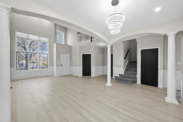 unfurnished living room with a wainscoted wall, light wood-type flooring, stairs, arched walkways, and ornate columns