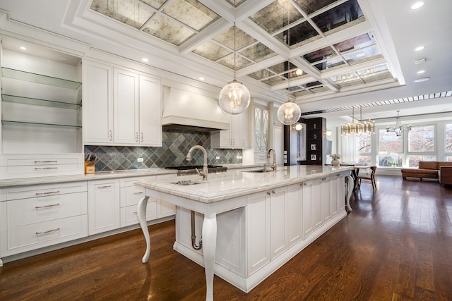 kitchen with coffered ceiling, a kitchen island with sink, premium range hood, white cabinetry, and pendant lighting