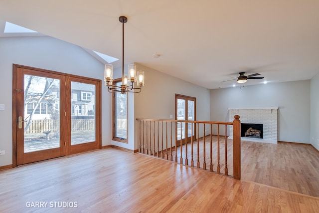 unfurnished living room featuring ceiling fan with notable chandelier, a brick fireplace, wood finished floors, and baseboards