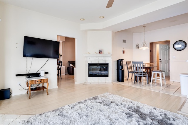 living area with ceiling fan, light wood finished floors, a tiled fireplace, and visible vents