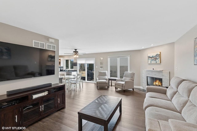 living room featuring a warm lit fireplace, visible vents, ceiling fan, and wood finished floors