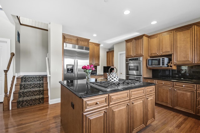 kitchen featuring built in appliances, brown cabinetry, dark stone counters, and a center island with sink