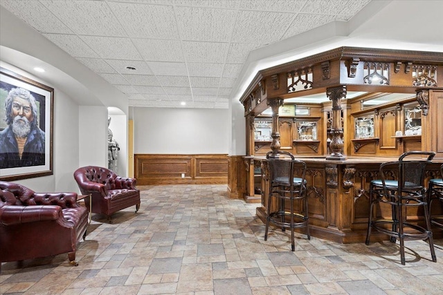 bar featuring arched walkways, a paneled ceiling, indoor bar, wainscoting, and stone finish floor