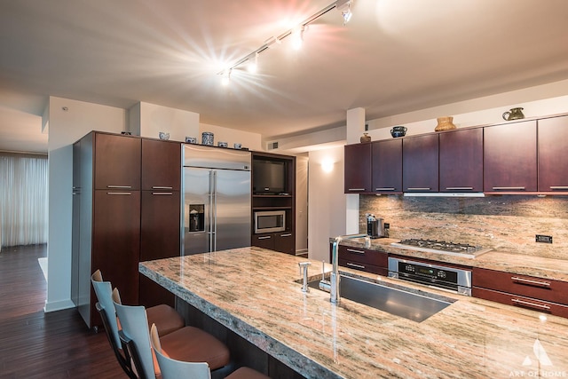 kitchen featuring tasteful backsplash, dark wood finished floors, built in appliances, a breakfast bar area, and a sink