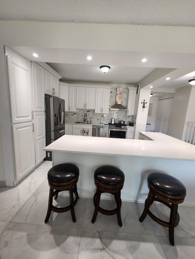 kitchen with a breakfast bar, marble finish floor, white cabinets, wall chimney range hood, and a peninsula