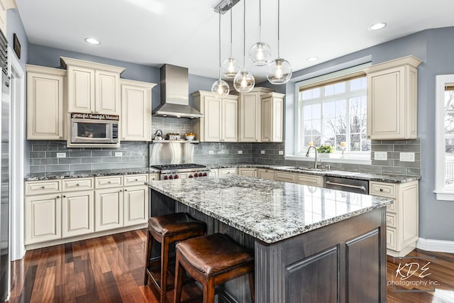 kitchen with cream cabinetry, appliances with stainless steel finishes, a kitchen island, wall chimney range hood, and a sink