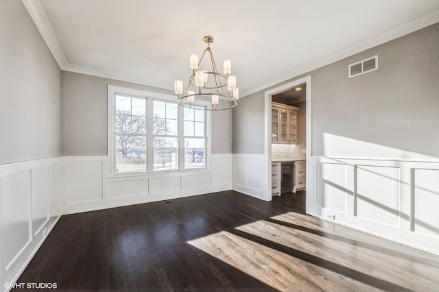 unfurnished dining area with a notable chandelier, dark wood-type flooring, visible vents, ornamental molding, and wainscoting
