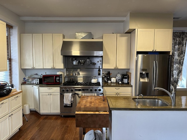 kitchen featuring dark countertops, dark wood-style floors, wall chimney exhaust hood, appliances with stainless steel finishes, and a sink