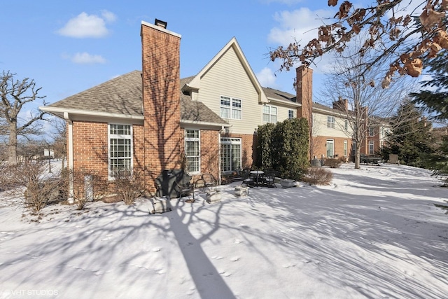 snow covered rear of property featuring brick siding, a chimney, and a shingled roof