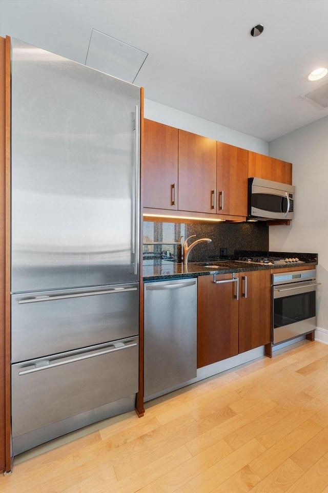 kitchen featuring backsplash, dark stone countertops, light wood-style floors, brown cabinetry, and stainless steel appliances