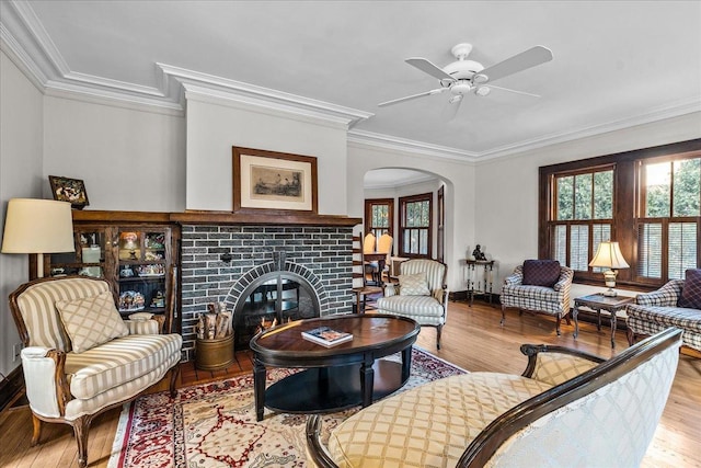 living room with a brick fireplace, ornamental molding, wood finished floors, arched walkways, and a ceiling fan