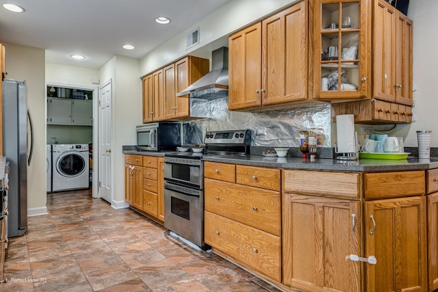 kitchen with dark countertops, glass insert cabinets, appliances with stainless steel finishes, wall chimney range hood, and washer / dryer