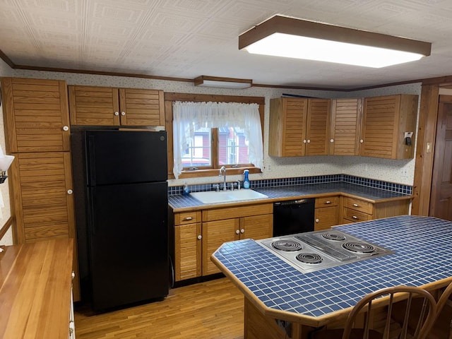 kitchen with a sink, tile counters, black appliances, brown cabinetry, and wallpapered walls