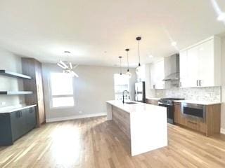 kitchen featuring white cabinets, an island with sink, wall chimney exhaust hood, stainless steel appliances, and light countertops