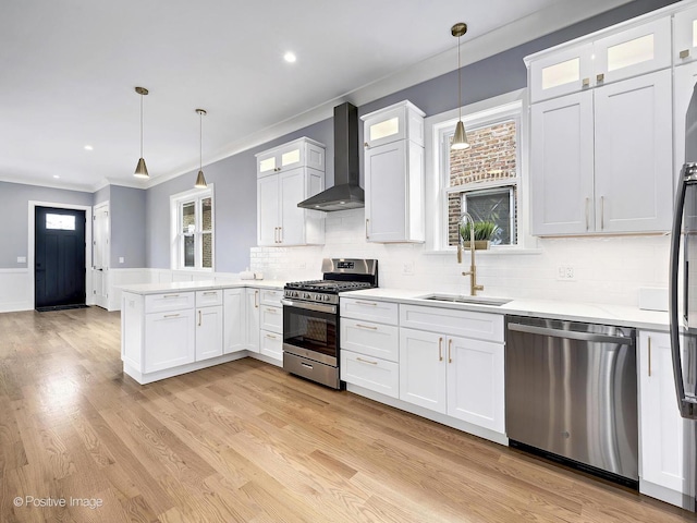 kitchen featuring glass insert cabinets, stainless steel appliances, light countertops, wall chimney range hood, and a sink