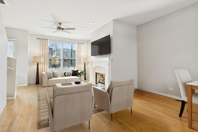 living room featuring light wood-style flooring, baseboards, ceiling fan, and a lit fireplace