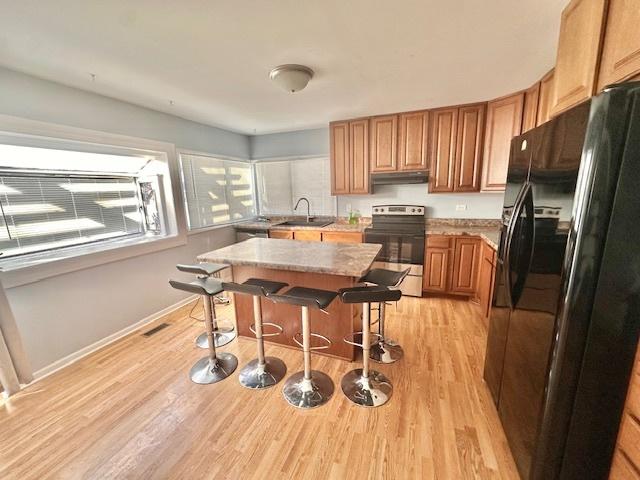 kitchen featuring stainless steel electric stove, light wood-type flooring, under cabinet range hood, a kitchen bar, and black fridge