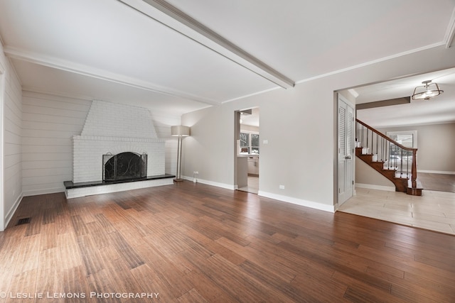 unfurnished living room with beam ceiling, a brick fireplace, wood finished floors, baseboards, and stairs