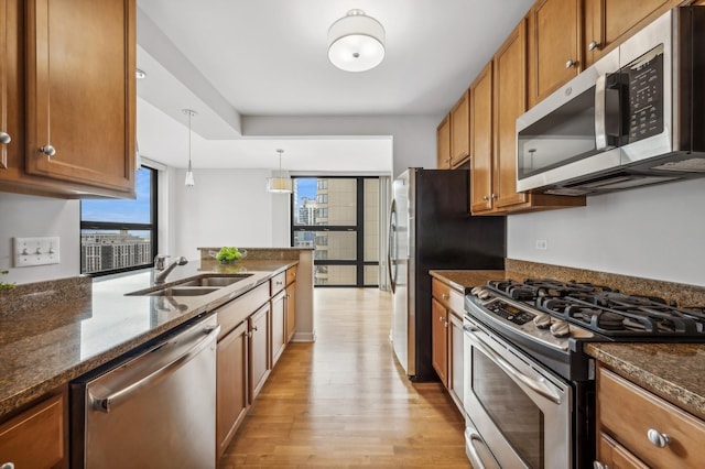 kitchen featuring brown cabinetry, appliances with stainless steel finishes, dark stone countertops, hanging light fixtures, and a sink