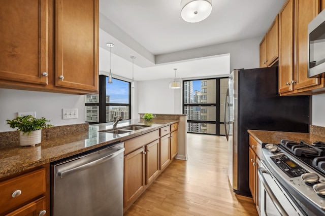 kitchen featuring a sink, appliances with stainless steel finishes, brown cabinets, dark stone counters, and decorative light fixtures