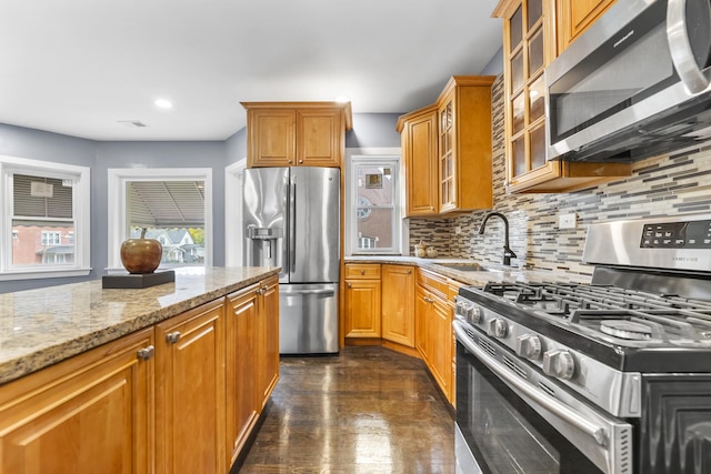 kitchen with glass insert cabinets, appliances with stainless steel finishes, a sink, light stone countertops, and backsplash