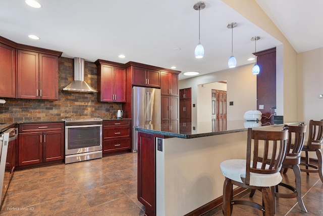 kitchen with a breakfast bar area, stainless steel appliances, wall chimney range hood, reddish brown cabinets, and decorative light fixtures