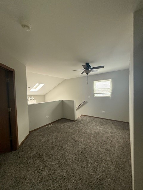 spare room featuring ceiling fan, dark colored carpet, lofted ceiling with skylight, and baseboards