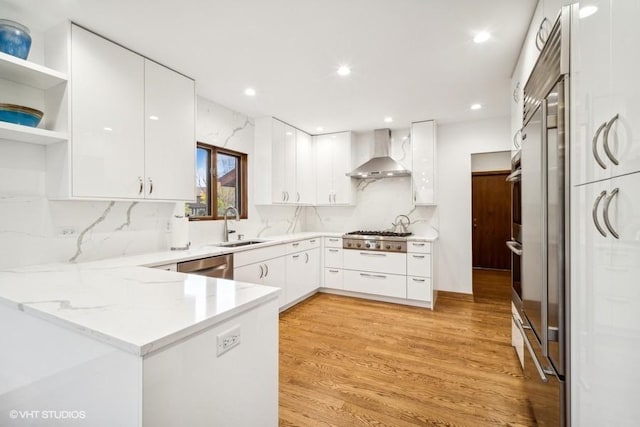 kitchen featuring open shelves, a peninsula, a sink, appliances with stainless steel finishes, and wall chimney exhaust hood