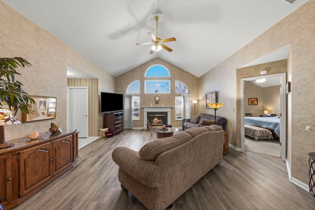 living room featuring ceiling fan, wood finished floors, high vaulted ceiling, a warm lit fireplace, and baseboards