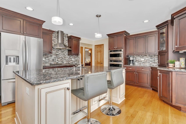 kitchen featuring wall chimney range hood, dark stone countertops, stainless steel appliances, and light wood-style floors