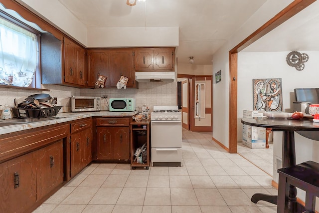 kitchen with white gas stove, light tile patterned flooring, under cabinet range hood, light countertops, and decorative backsplash