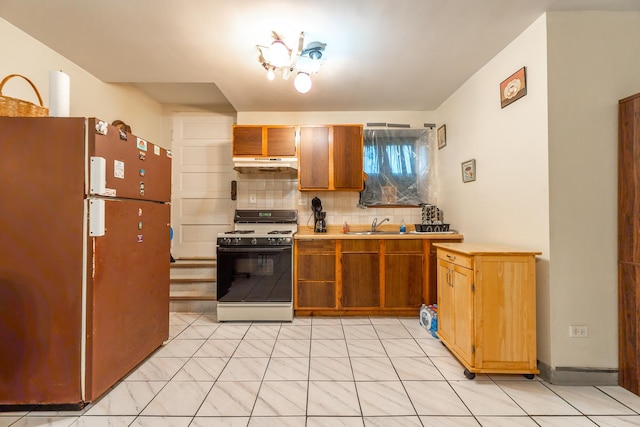 kitchen featuring under cabinet range hood, a sink, backsplash, freestanding refrigerator, and gas range