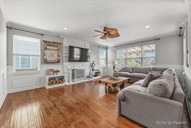 living area with a ceiling fan, hardwood / wood-style flooring, a wainscoted wall, ornamental molding, and a stone fireplace