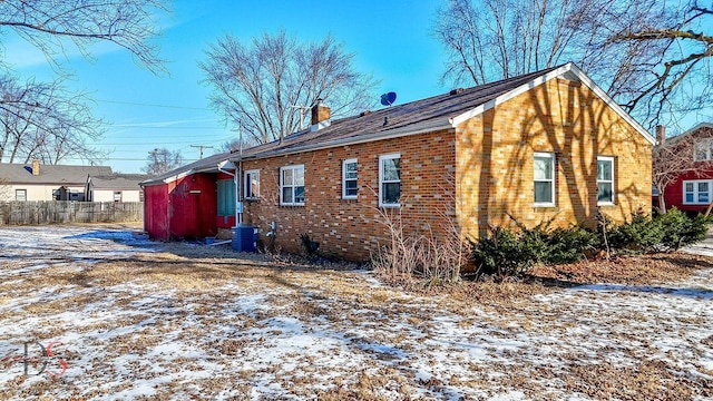 snow covered house with brick siding, a chimney, central AC unit, and fence