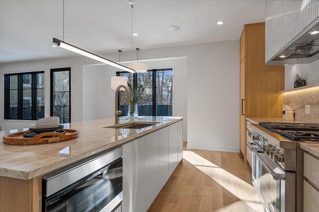 kitchen with range with two ovens, light stone counters, a sink, light wood-style floors, and wall chimney exhaust hood