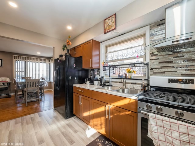 kitchen featuring a sink, wall chimney range hood, brown cabinets, black refrigerator with ice dispenser, and gas stove