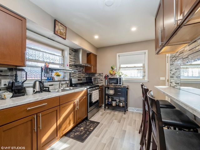 kitchen featuring stainless steel appliances, backsplash, brown cabinetry, a sink, and wall chimney exhaust hood