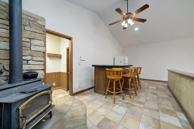 bar with a wainscoted wall, a dry bar, a ceiling fan, a wood stove, and vaulted ceiling