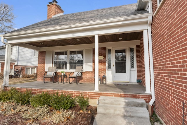 entrance to property featuring covered porch, brick siding, roof with shingles, and a chimney