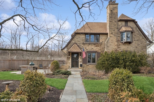 tudor home featuring roof with shingles, fence, a chimney, and brick siding