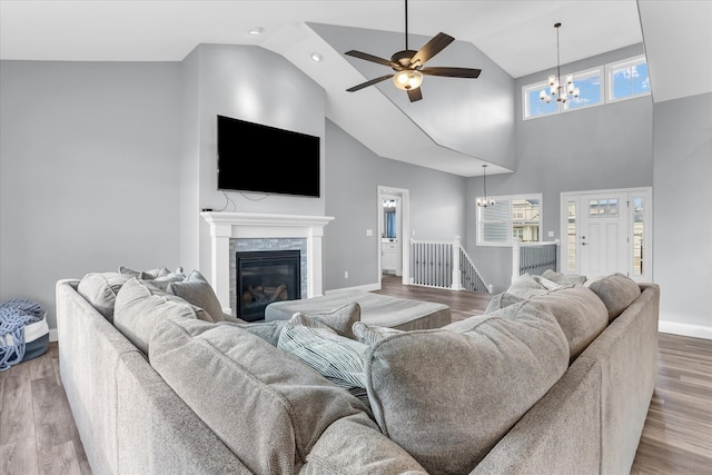 living room featuring wood finished floors, baseboards, high vaulted ceiling, a stone fireplace, and ceiling fan with notable chandelier